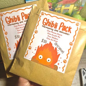 K&ouml;nnte beinhalten: Zwei braune Papiert&uuml;ten mit orangefarbenen und wei&szlig;en "Ghibli Pack"-Etiketten. Die Etiketten zeigen den Text "Ghibli Pack" und eine Artikelliste. Ein Aufkleber hat eine Cartoon-Feuer-Grafik. Eine Hand h&auml;lt die T&uuml;ten.