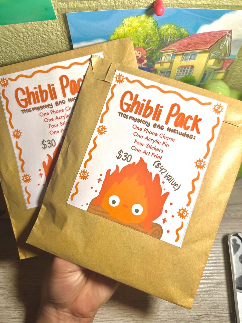 K&ouml;nnte beinhalten: Zwei braune Papiert&uuml;ten mit orangefarbenen und wei&szlig;en "Ghibli Pack"-Etiketten. Die Etiketten zeigen den Text "Ghibli Pack" und eine Artikelliste. Ein Aufkleber hat eine Cartoon-Feuer-Grafik. Eine Hand h&auml;lt die T&uuml;ten.