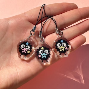 May include: Three clear acrylic charms with black, cartoon-like characters with star and bow tie details. Each charm has a black cord and silver-colored metal clasp. The charms are displayed against a pink background.