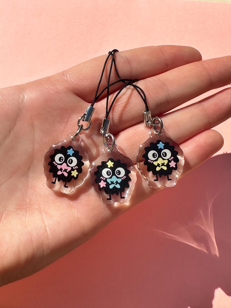May include: Three clear acrylic charms with black, cartoon-like characters with star and bow tie details. Each charm has a black cord and silver-colored metal clasp. The charms are displayed against a pink background.