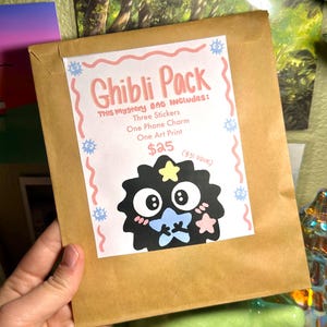 May include: A brown paper bag labeled "Ghibli Pack" with a cartoon character illustration. The bag includes three stickers, a phone charm, and an art print. The text on the bag reads "THIS Mystery BAG INCLUDES: Three Stickers, One Phone Charm, One Art Print."