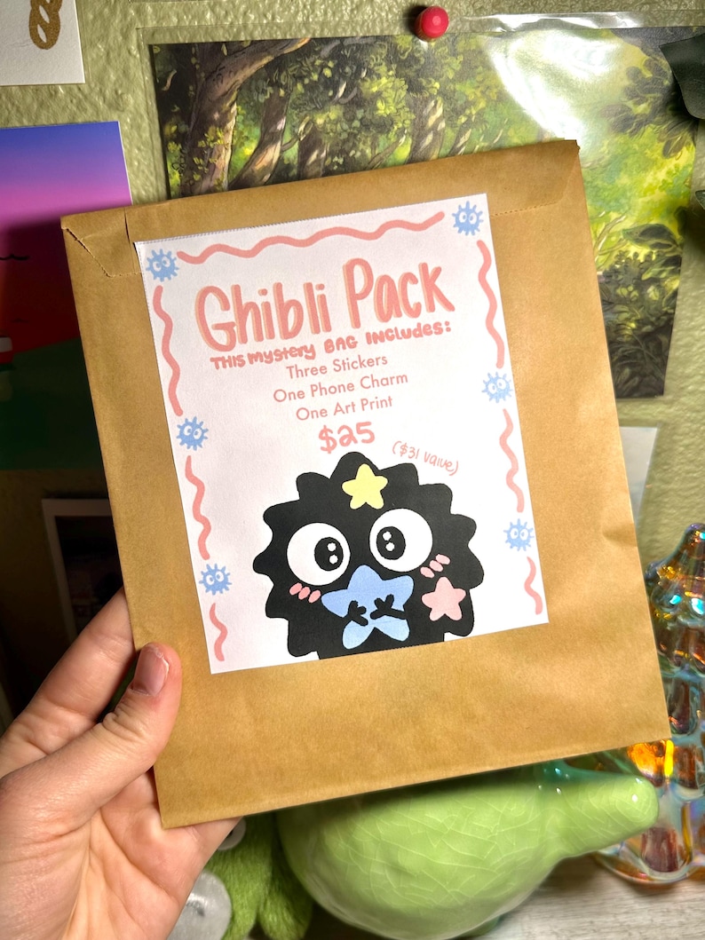 May include: A brown paper bag labeled "Ghibli Pack" with a cartoon character illustration. The bag includes three stickers, a phone charm, and an art print. The text on the bag reads "THIS Mystery BAG INCLUDES: Three Stickers, One Phone Charm, One Art Print."
