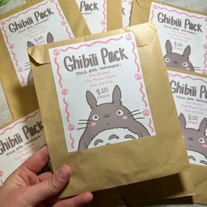Pu&ograve; includere: Diverse buste di carta marrone con un'etichetta bianca che recita "Ghibili Pack" con un'illustrazione di un personaggio dei cartoni animati. Ogni busta include due adesivi, un ciondolo per telefono e una stampa artistica.
