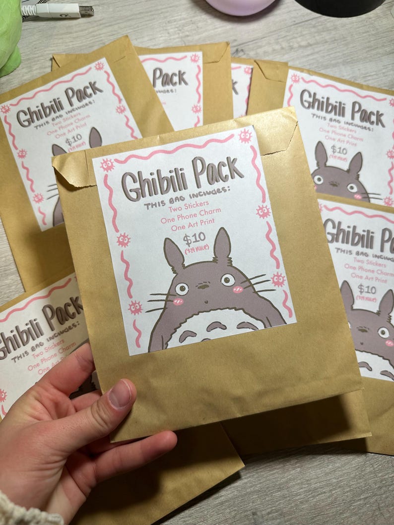 Pu&ograve; includere: Diverse buste di carta marrone con un'etichetta bianca che recita "Ghibili Pack" con un'illustrazione di un personaggio dei cartoni animati. Ogni busta include due adesivi, un ciondolo per telefono e una stampa artistica.