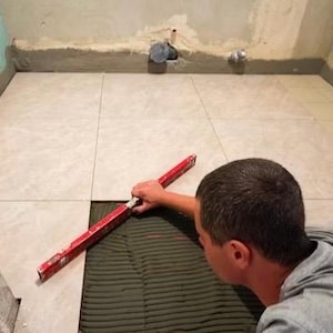 Może przedstawiać: Osoba używa czerwonej poziomicy, aby sprawdzić wyrównanie dużych, kwadratowych płytek podłogowych w kolorze beżowym z marmurowym wzorem. Płytki są układane na warstwie ciemnoszarej zaprawy klejącej na podłożu w niewykończonym pomieszczeniu.