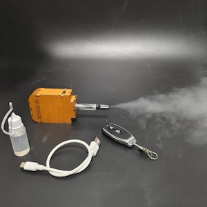 May include: A rectangular wooden device emitting a plume of white vapor from a metallic nozzle, accompanied by a small clear plastic bottle with a white cap, a white USB-C charging cable, and a black remote control featuring buttons labeled A and B.
