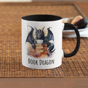 Puede incluir: Una taza de cerámica blanca con un borde y un asa negros. La taza presenta una ilustración de acuarela de un dragón negro sentado en una pila de libros y sosteniendo una taza de té. El texto "BOOK DRAGON" está impreso en negro en la parte inferior de la taza.