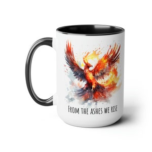 May include: White ceramic coffee mug with a black rim and handle. The mug features a watercolor illustration of a phoenix rising from flames with the text "From the ashes we rise".