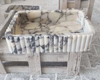 May include: A white marble sink with a wavy, fluted edge. The sink is sitting on a wooden pallet.
