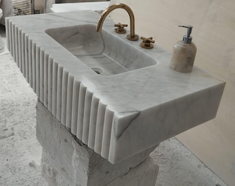 Puede incluir: Vanidad de baño de mármol blanco con un borde acanalado, un lavabo de porcelana blanca, un grifo dorado y un dispensador de jabón dorado.