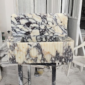 Può includere: Un lavandino rettangolare in marmo con alzatina. Il lavandino ha una base color crema con venature grigio scuro e nere. La parte anteriore del lavandino presenta delle scanalature verticali. L'alzatina ha un motivo simile. Il lavandino è su un supporto in metallo nero.