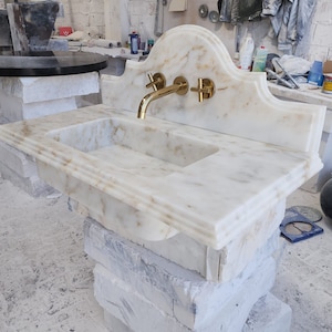 Op de afbeelding: Een wastafel van wit marmer met goudkleurige metalen kranen, inclusief een wandkraan en twee kruisgrepen. De wastafel heeft een rechthoekige bak en een decoratieve, gebogen achterwand met afgeschuinde randen en natuurlijke aders.