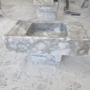 May include: A white marble sink with a rectangular shape and a single basin. The sink is sitting on a concrete base.
