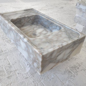 May include: A white marble sink with gray veining. The sink is rectangular and has a square shape. The sink is made of natural stone and has a smooth, polished finish.