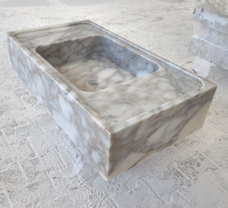 May include: A white marble sink with gray veining. The sink is rectangular and has a square shape. The sink is made of natural stone and has a smooth, polished finish.