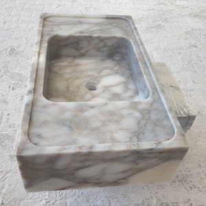 May include: A white marble bathroom sink with a rectangular shape and a single drain hole. The sink has a smooth, polished surface with gray and brown veining.