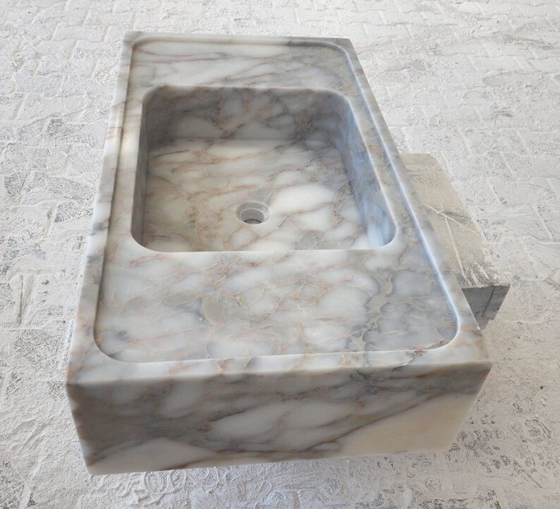 May include: A white marble bathroom sink with a rectangular shape and a single drain hole. The sink has a smooth, polished surface with gray and brown veining.