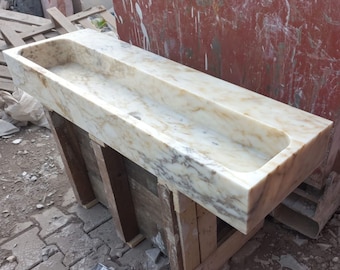 May include: A white marble sink with a rectangular shape and a long, narrow basin. The sink is resting on wooden supports.