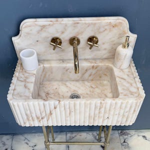 Puede incluir: Un lavabo de mármol con grifo y accesorios dorados. El lavabo tiene una cubeta rectangular y un borde festoneado. Un dispensador de jabón y un portacepillos a juego están sobre la encimera. El lavabo está sostenido por patas doradas.