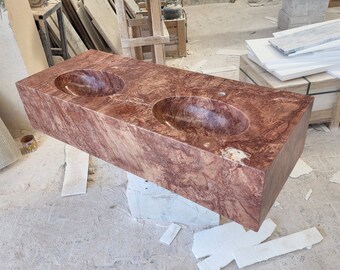 May include: A double sink bathroom vanity made of red marble with a natural veined pattern. The sink is made of a single piece of marble with a smooth, polished finish.