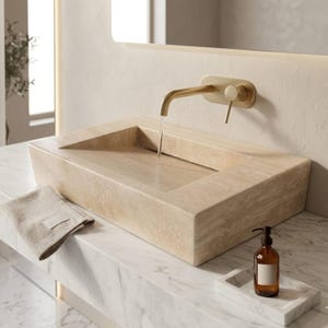 Può includere: Un lavabo rettangolare in pietra beige con un rubinetto dorato. L'acqua scorre nel lavandino. Un piccolo asciugamano beige e una bottiglia marrone con un'etichetta bianca sono sul piano di lavoro in marmo.
