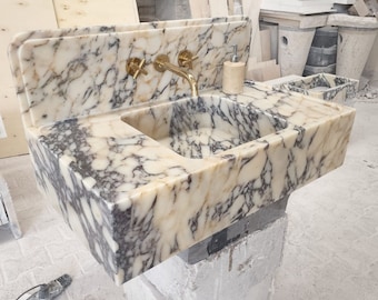May include: A white marble bathroom sink with a gold faucet and soap dispenser. The sink is mounted on a wall and has a backsplash. The sink is made of natural stone and has a unique veining pattern.