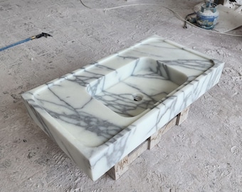 Puede incluir: Un lavabo rectangular de mármol con una pila y un desagüe integrados. El lavabo presenta una base blanca con vetas grises. El lavabo está colocado sobre una estructura de soporte de madera.