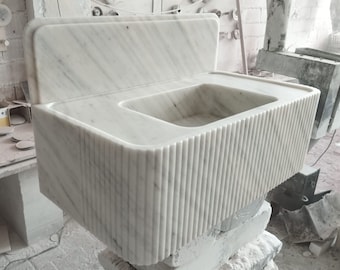 May include: A white marble sink with a raised backsplash and a fluted front. The sink is sitting on a stack of blocks.