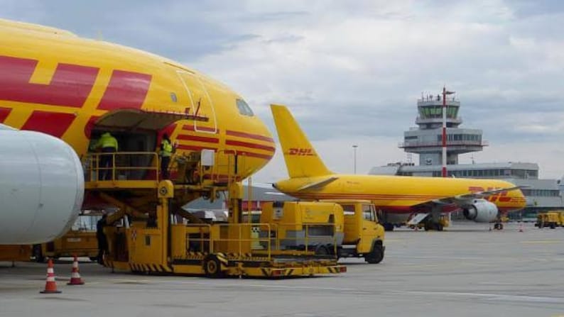May include: Two yellow DHL cargo planes are parked at an airport. The plane in the foreground is being loaded with cargo. The plane in the background is a Boeing 757-200F.