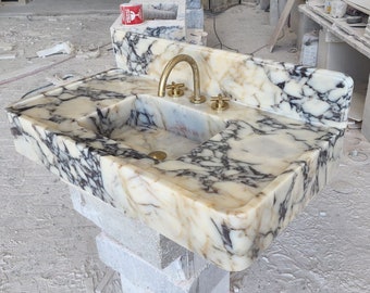 Puede incluir: Un lavabo de baño de mármol blanco con vetas negras. El lavabo tiene una forma rectangular y un grifo simple con dos manijas. El lavabo está montado en una encimera de mármol blanco.