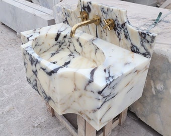 Può includere: Un lavandino in marmo con un motivo venato crema e nero. Il lavandino ha un bacino arrotondato e un alzatina decorativa. È dotato di un rubinetto dorato e maniglie a forma di croce. Il lavandino è montato su un supporto in legno.
