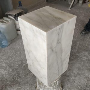 May include: A white marble pedestal with a square top and a smooth, polished finish. The pedestal is sitting on a gray concrete floor.