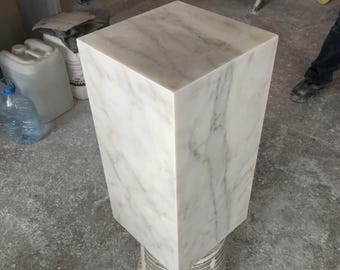Könnte beinhalten: Ein weißer Marmorsockel mit quadratischer Oberseite und glatter, polierter Oberfläche. Der Sockel steht auf einem grauen Betonboden.