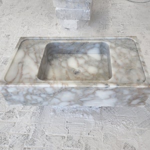 May include: A white marble sink with a rectangular shape and a square basin. The sink has a smooth, polished surface with a subtle veining pattern.