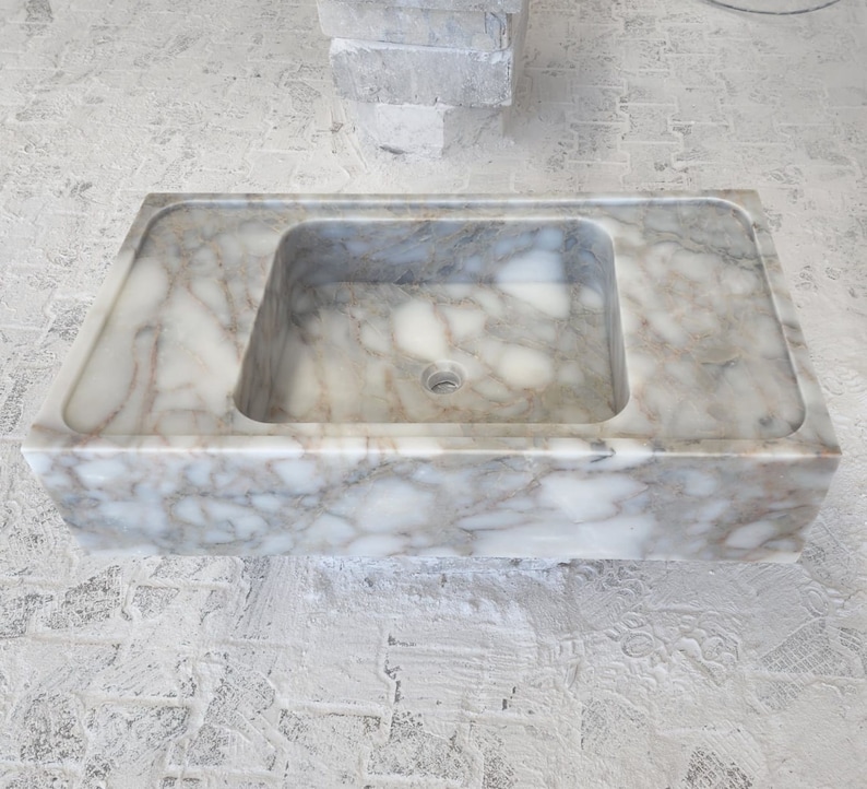 May include: A white marble sink with a rectangular shape and a square basin. The sink has a smooth, polished surface with a subtle veining pattern.