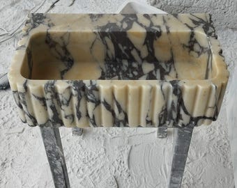 May include: A white marble sink with black veining, featuring a fluted edge and a rectangular shape. The sink is mounted on a metal stand with two legs.