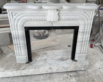 May include: A white marble fireplace with a black rectangular opening. The fireplace features ornate carvings, including a central floral design and geometric patterns. The structure is set on a matching marble base.