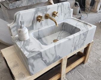 Op de afbeelding: Een witte marmeren wastafel met een gouden kraan en een zeepdispenser. De wastafel is gemonteerd op een houten standaard.