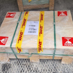 May include: A wooden crate with a yellow and black DHL tape, wrapped in plastic and secured with green straps. The crate has red and white "DİKKAT KIRILIR" (Turkish for "Caution Fragile") stickers with a glass symbol.