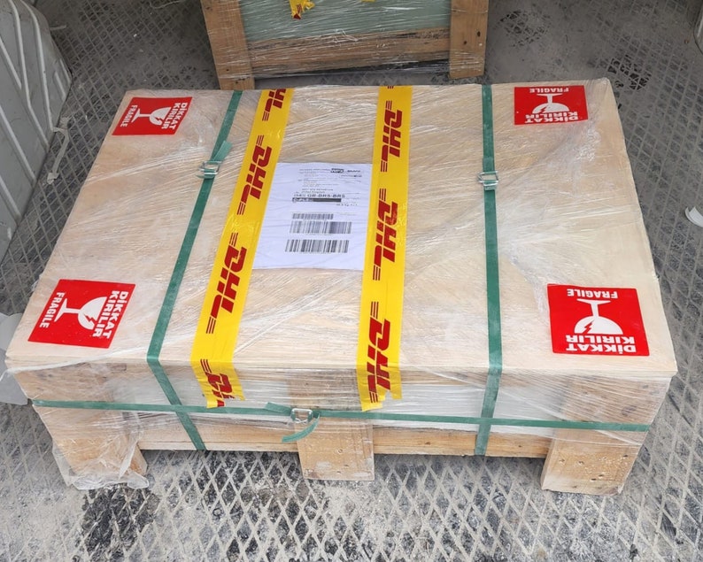 May include: A wooden crate with a yellow and black DHL tape, wrapped in plastic and secured with green straps. The crate has red and white "DİKKAT KIRILIR" (Turkish for "Caution Fragile") stickers with a glass symbol.