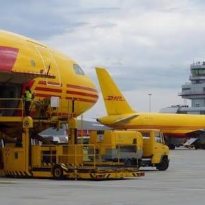 May include: A yellow and red DHL cargo plane is being loaded with cargo by a yellow loading vehicle. The plane is parked on the tarmac at an airport.