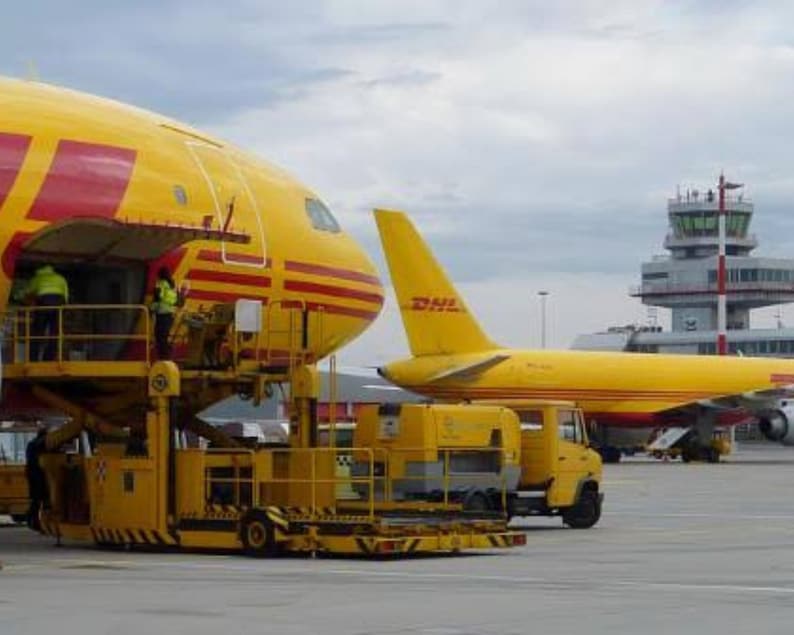 May include: A yellow and red DHL cargo plane is being loaded with cargo by a yellow loading vehicle. The plane is parked on the tarmac at an airport.