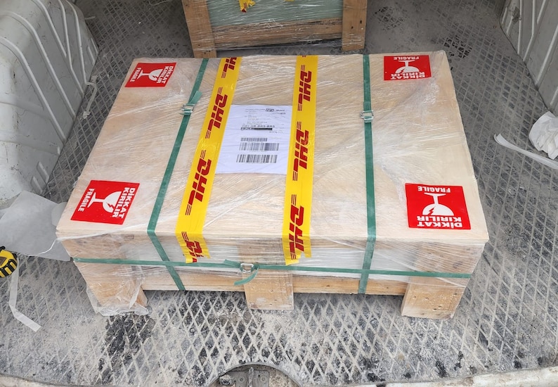 May include: A wooden crate wrapped in plastic and secured with yellow DHL tape. The crate has red and white "DİKKAT KIRILIR FRAGILE" stickers on each side.