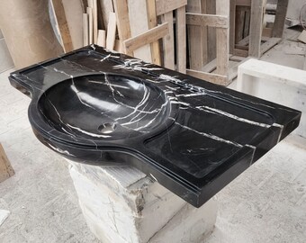 May include: Black marble bathroom sink with a white veined pattern. The sink is oval shaped and has a raised edge around the basin. The sink is mounted on a white countertop.