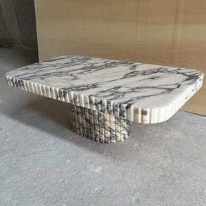 Könnte beinhalten: Ein rechteckiger Couchtisch aus Marmor mit einer weiß-grau geäderten Platte. Der Tisch hat eine geriffelte Kante und einen zylindrischen Sockel, ebenfalls aus Marmor. Der Tisch ist ein modernes Design.
