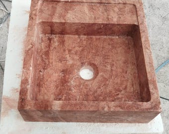 May include: A square, red-brown stone sink with a single drain hole. The sink has a raised edge and a slightly recessed area around the drain.