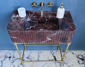 Puede incluir: Un lavabo de mármol rojo con vetas blancas, sobre un soporte de metal dorado. El lavabo tiene una cubeta rectangular y un exterior acanalado. Un grifo dorado y dos accesorios de pared están encima del lavabo. Una taza y un dispensador de jabón blancos están en la superficie.
