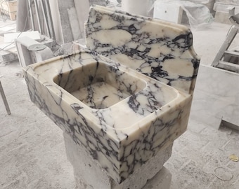 May include: A white marble sink with black veining. The sink has a raised backsplash and is mounted on a concrete base.