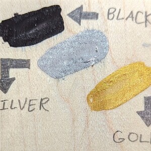 May include: Three paint swatches on a wooden surface. The swatches are labelled "BLACK", "SILVER", and "GOLD" with arrows pointing to each swatch.