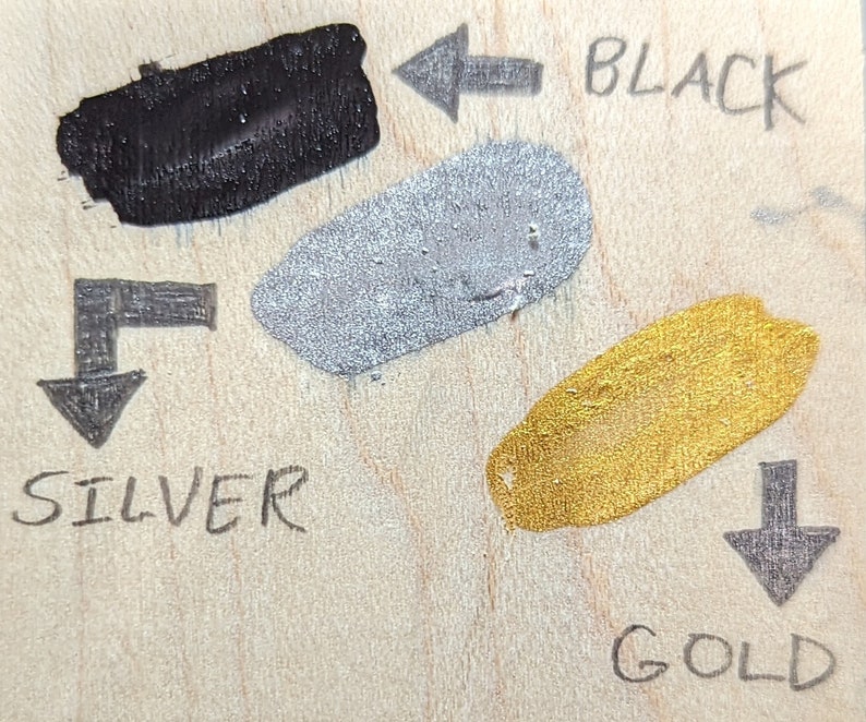 May include: Three paint swatches on a wooden surface. The swatches are labelled "BLACK", "SILVER", and "GOLD" with arrows pointing to each swatch.